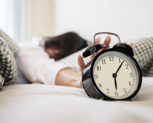 Sleepy woman reaching  holding the alarm clock in the morning with late wake up - every day life at home concept